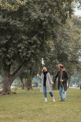 Fototapeta premium full length of positive redhead man and cheerful woman holding coffee to go while walking in park.