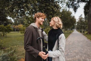 happy blonde woman and cheerful redhead man with umbrella holding hands in park.