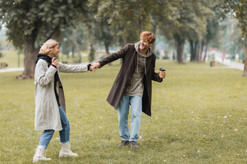 Naklejka na ściany i meble full length of joyful man in coat holding hands with smiling girlfriend in autumnal park.