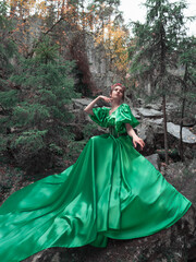 Fototapeta premium A beautiful woman in a long green dress with a corset and a decorative crown of branches and flowers on her head. Forest nymph on the background of rocks. Full length portrait.