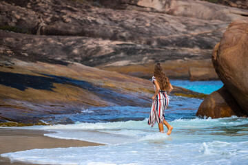 a long-haired girl in a long dress soaks her feet in the water on a paradise beach in australia with massive rocks in the background; hellfire bay in cape le grand national park in australia for