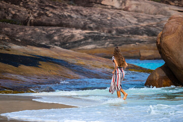 a long-haired girl in a long dress soaks her feet in the water on a paradise beach in australia with massive rocks in the background; hellfire bay in cape le grand national park in australia for