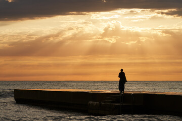 A lonely female figure on the pier at sunset. The last rays of the sun are breaking through a gap in the clouds. Copy  space.