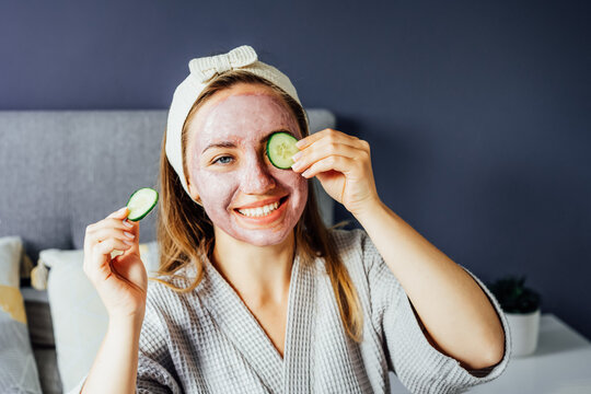 A Young Smiling Woman With Pink Clay Facial Mask Holds Cucumber Slices Making A Refreshing Eye Mask In Bedroom. Natural Cosmetic Procedures For Skin Care At Home. Beauty Self-care. Selective Focus