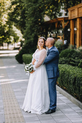 An adult stylish groom in a blue suit and a beautiful smiling bride in a white dress are hugging on a city street in a park. Wedding photography, portrait.