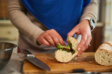Details: Woman's hands put greens and organic fresh vegetables in the bread, making crusty sandwiches with veggies in the home kitchen, preparing a delicious healthy snack