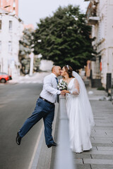 Fototapeta na wymiar A stylish adult groom and a beautiful smiling bride in a white dress are hugging, kissing on the street in the city near the railings. Wedding photography, portrait.
