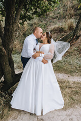 A stylish adult groom and a beautiful, young, smiling bride in a white dress are hugging, standing in nature near a tree. Close-up wedding photography, portrait.