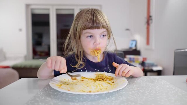 Hungry child eats pasta greedily. Little girl with disheveled hair and dirty face finishes her dinner. Kid enjoying eating. Baby's face and hands covered in tomato paste. Toddler has a big appetite.
