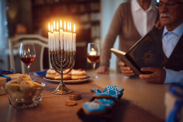 Lit candles in menorah with Jewish couple reading Hebrew bible in background. - obrazy, fototapety, plakaty