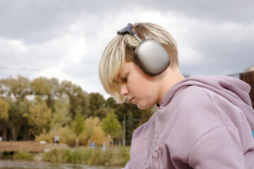 Sad lonely pensive teenager boy listening to music on wireless headphones walking outside in the park. Teenage issues and problems concept