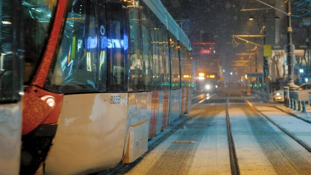 Snow flakes falling in city street at night, tram moving along rails, blizzard or snowstorm in Istambul. Urban cityscape background, city blurred lights, walking people 