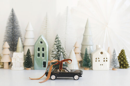 Holidays Are Coming! Stylish Little Car Carrying Christmas Tree On Background Of Christmas Miniature Snowy Village. Merry Christmas! Festive Winter Scene On White Table, Holiday Banner