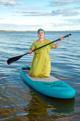 A woman in a green dress and a scarf on her knees on a sup board against a clear blue sky.