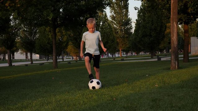 An active eight-year-old boy trains in a city park with a soccer ball in the summer at sunset.