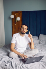Fototapeta premium man in a white T-shirt, a freelancer with glasses, sits on the bed at home with a laptop and works remotely in the morning. suddenly came a brilliant idea