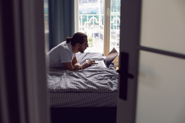 man in a white T-shirt, a freelancer with glasses, lie on the bed at home with a laptop and works remotely in the morning. lies in front of a large window.