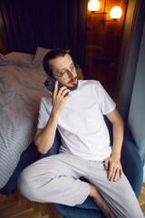 portrait of a bearded man in glasses, white T-shirt and house pants sitting on a chair by the window with a phone. by the bed.