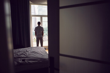 man with long hair, beard and glasses stands at a large window in an apartment with a view of a skyscraper in the morning. the guy is dressed in a white T-shirt and sweatpants barefoot.