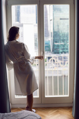 portrait of a beautiful brunette woman in a bathrobe stands at a large window in an apartment opposite a skyscraper in the morning. - obrazy, fototapety, plakaty