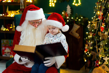 Fototapeta na wymiar Santa Clause reads a book to a little cute boy while sitting near Christmas tree. New Year concept 