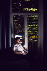 freelance man in glasses working in shorts and a T-shirt sits at the window on the floor in front of a skyscraper at night.