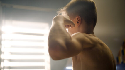 Obraz na płótnie Canvas Young boy checks his biceps after dumbbells curls in front of the mirror. Closeup of his biceps and abs. Eaxmines upper body after exercuse. Weight training, fitness, physical activity.