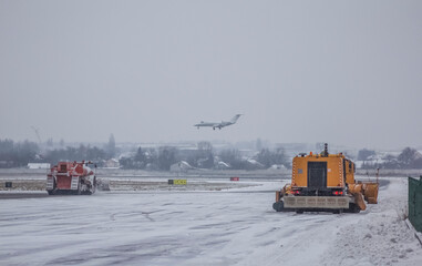 A business jet is landing against the backdrop of airport snowplows against a cloudy sky in winter
