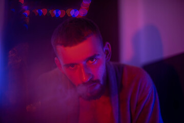 A brutal man with a beard sits in a bar, smokes and exhales smoke. A guy looks at the camera in purple light.