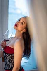 A female goddess in a red dress with a corset holds a glass of wine in her hand. Looks fascinated at the camera while standing near the window.