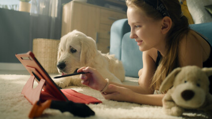 Young girl playing with dog on tablet, lying on mild carpet, showing gaming animations, smiling and having fun, spending leisure time at home. Golden retriever.