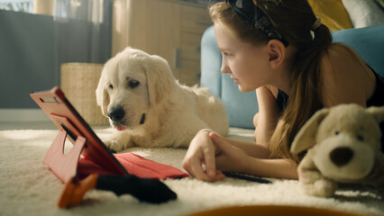 Young girl playing with dog on tablet, lying on mild carpet, showing gaming animations, smiling and...