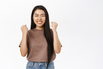 Yes, achievement. Happy asian girl wins, celebrates success and victory, triumphs, makes fist pumps, stands over white background