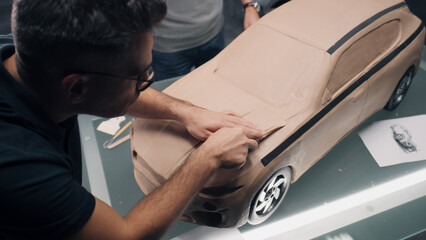 Two male automotive designers sculpting futuristic plasticine clay model of car with rake. Future...