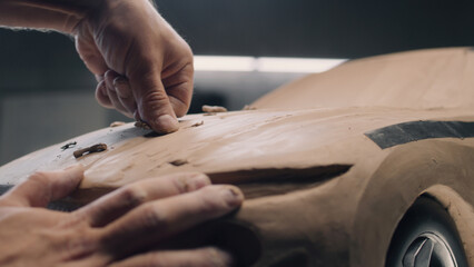 Close-up shot of male automotive designer sculpting eco friendly electric car model from plasticine clay with rake. Future design of new generation electric car. Working in modern car design studio.