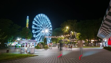 Ferris wheel in amusement park beautifully illuminated in the evening, spinning in motion timelapse. People walking around and having fun among carousels and attractions.