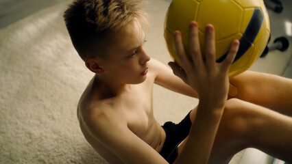 Young enthusiastic boy training abs holds a football while exercising alone indoors. Prepares before the soccer match. Sport and active lifestyle concept.
