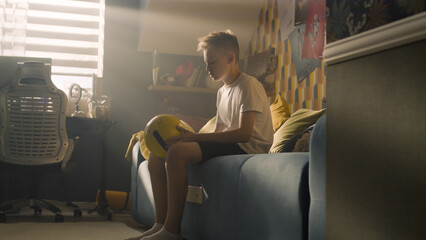 A teenager boy plays football while sitting on the bed alone in his room. Balancing the ball on his finger. Performing tricks. Sports, hobbies concept.