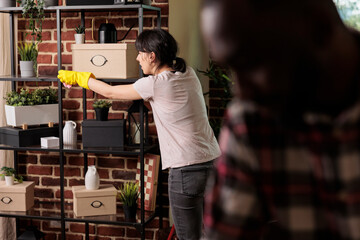 Woman wearing rubber gloves dusting shelves in urban style apartment, african american man helping...