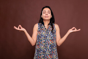 Relaxed indian woman meditating, showing om sign with fingers, front view studio medium shot. Serene person with closed eyes, calm lady with yoga symbol, meditation gesture, harmony concept