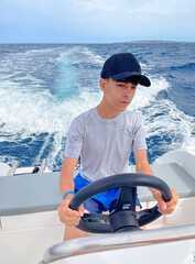 a young teenager, wearing a gray T-shirt and blue cap, enjoys crewing a sport boat leaving a trail...