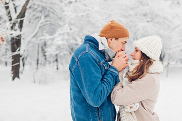 Man warming hands of woman with his breath in snowy cold winter park. Loving couple walking outdoors