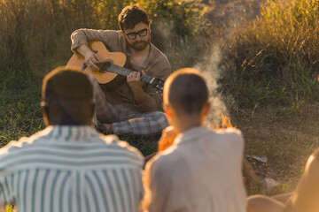 Fototapeta na wymiar Caucasian bearded buy in glasses sitting in front of his friends on the grass and playing acoustic guitar. Outdoor shot. Blurred people in the foreground. High quality photo