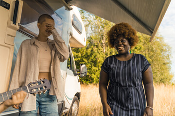 Acoustic guitar - live music during private party. Best friends - caucasian queer bald person and smiling Black woman in dark-blue dress - joking around and laughing in front of travel van. High - obrazy, fototapety, plakaty