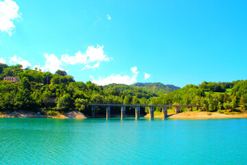 Tidy view of Gerosa Lake with a massive bridge suspended above the pure slightly rippled blue waters and reaching two hills chock full of vegetation and some buildings under a clear blue sky
