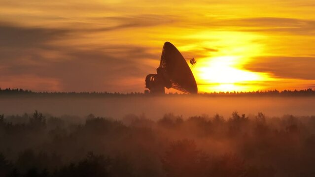 Aerial view. A giant radio telescope for space exploration in a foggy forest during a bloody dawn. Mysterious haze over the forest the sun's rays of dawn break through the red clouds.