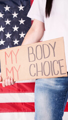 Young woman protester holds cardboard with My Body My Choice sign against USA flag on background....