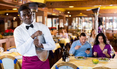 Portrait of upset tired adult african american waiter standing with empty serving tray in busy restaurant hall..