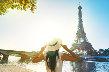 Young woman tourist in sun hat and white dress standing in front of Eiffel Tower in Paris at...