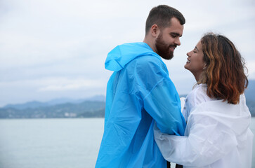 Young couple in raincoats enjoying time together under rain on beach, space for text